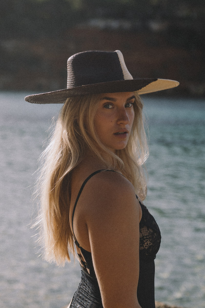 A woman in a body and black and white hat is posing for a shoot in front of the sea.