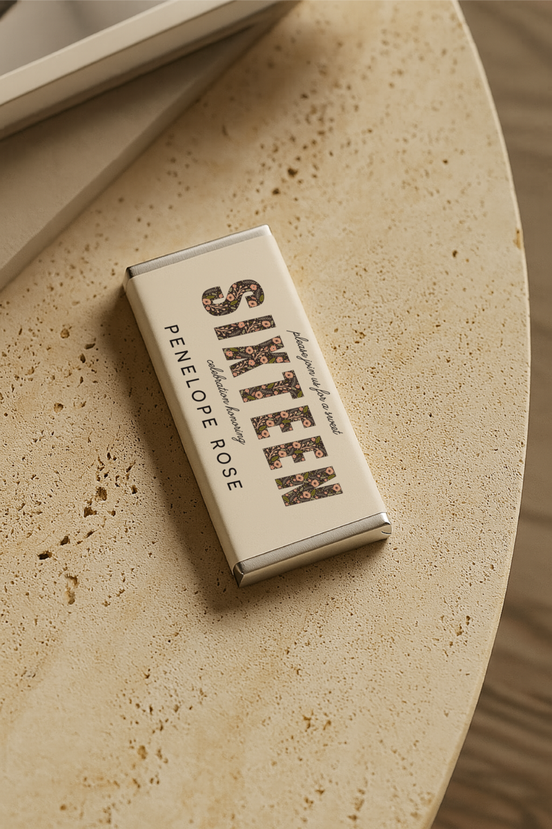 Personalised sweet sixteen chocolate bar with silver foil edges, floral typography, and custom name “Penelope Rose” on an ivory wrapper, styled on a stone surface.