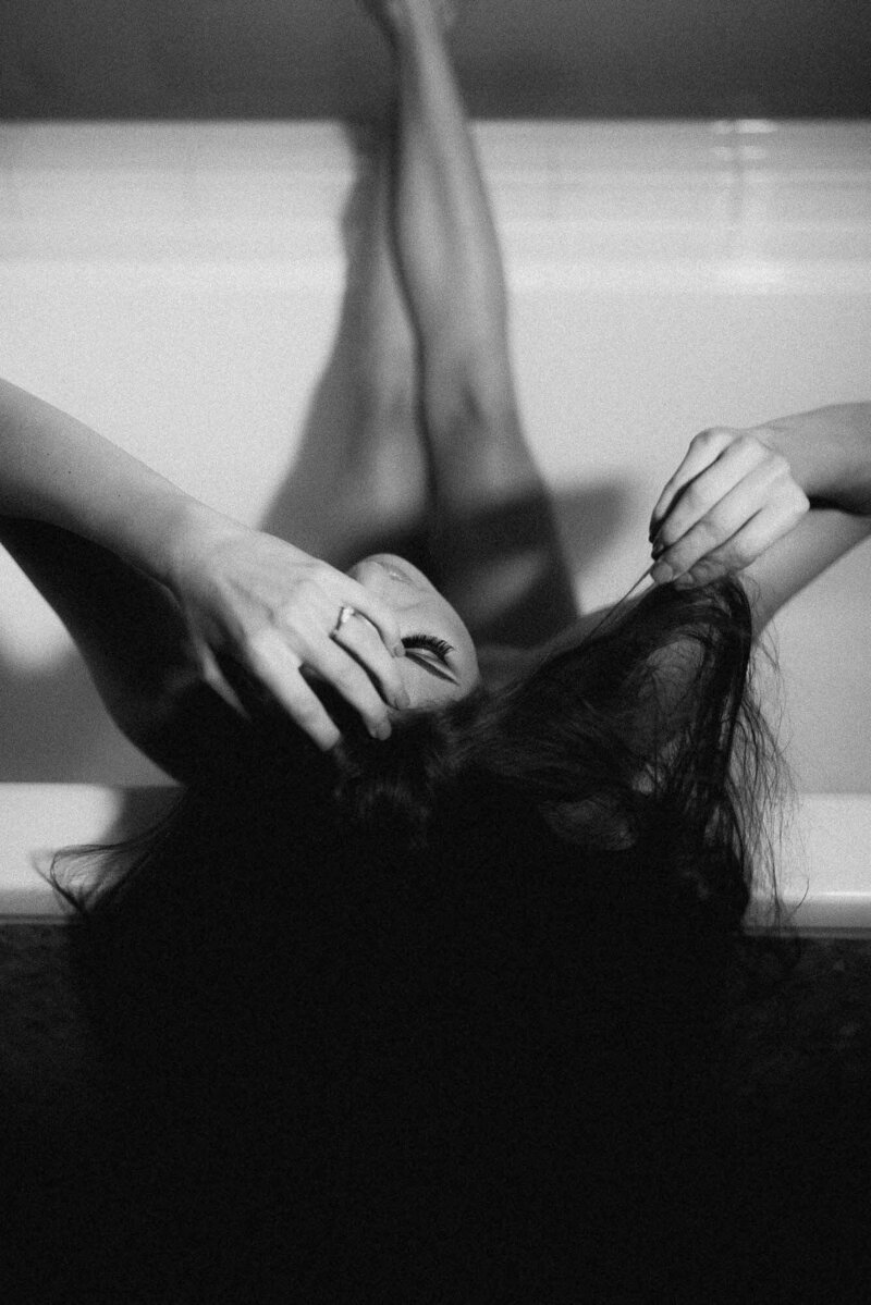 Black and white boudoir portrait of a woman in a bathtub with her legs extended, gently holding her hair; captured during a luxury boudoir session in Phoenix, Arizona.