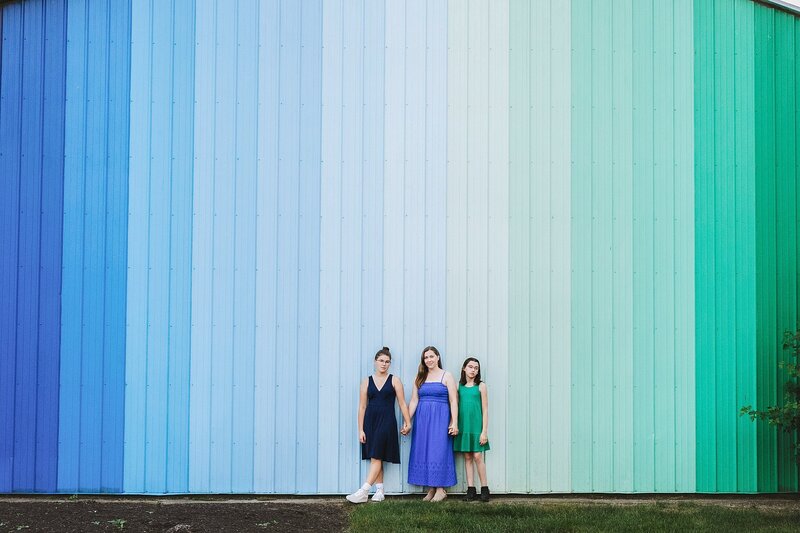 a-vibrant-family-photography-session-near-broad-ripple-indiana-featuring-a-mother-and-her-two-daughters-posing-against-a-striking-blue-to-green-gradient-mural-the-mother-wears-a-royal-blue-strapless-maxi-dress-one-daughter-wears-a-navy-blue-sleeveless-dress-with-white-sneakers-and-the-other-daughter-wears-an-emerald-green-sleeveless-dress-with-black-ankle-boots-captured-during-warm-weather-in-this-charming-urban-area-known-for-its-unique-murals-and-artistic-vibe-amanda-duncan-photography-specializes-in-natural-heartfelt-family-portraits-in-indianapolis-and-surrounding-areas







