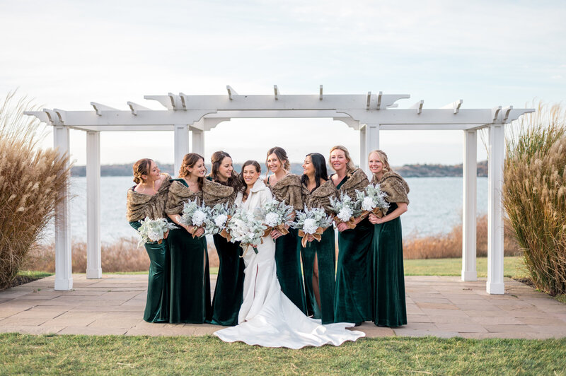 A bride and her bridesmaids in emerald green velvet dresses and fur shawls pose under a white pergola at Oceancliff, with sweeping water views behind them — perfect for an elegant winter wedding.