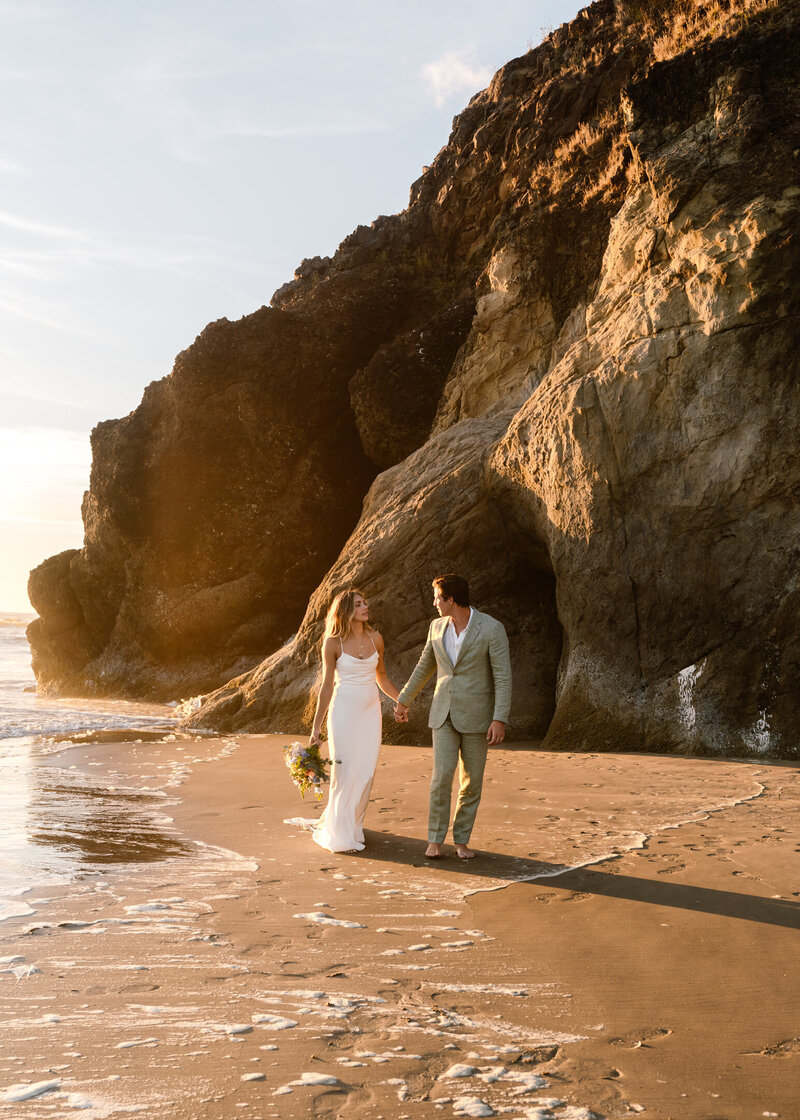 As the sun sets during their Oregon coast elopement, a couple, in their wedding attire walk on the beach. behind them is a large sea rock painted gold by the sun.