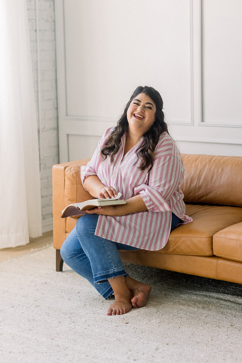 Marissa Martinez in a pink and white striped top and jeans holding her Bible and smiling while sitting on a brown leather couch