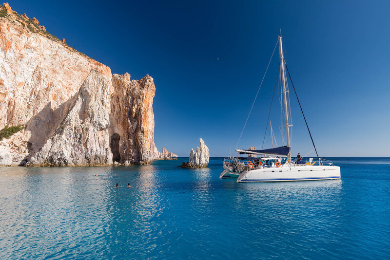 Sailboat anchored near white cliffs and clear blue water on a sunny day in a coastal bay.