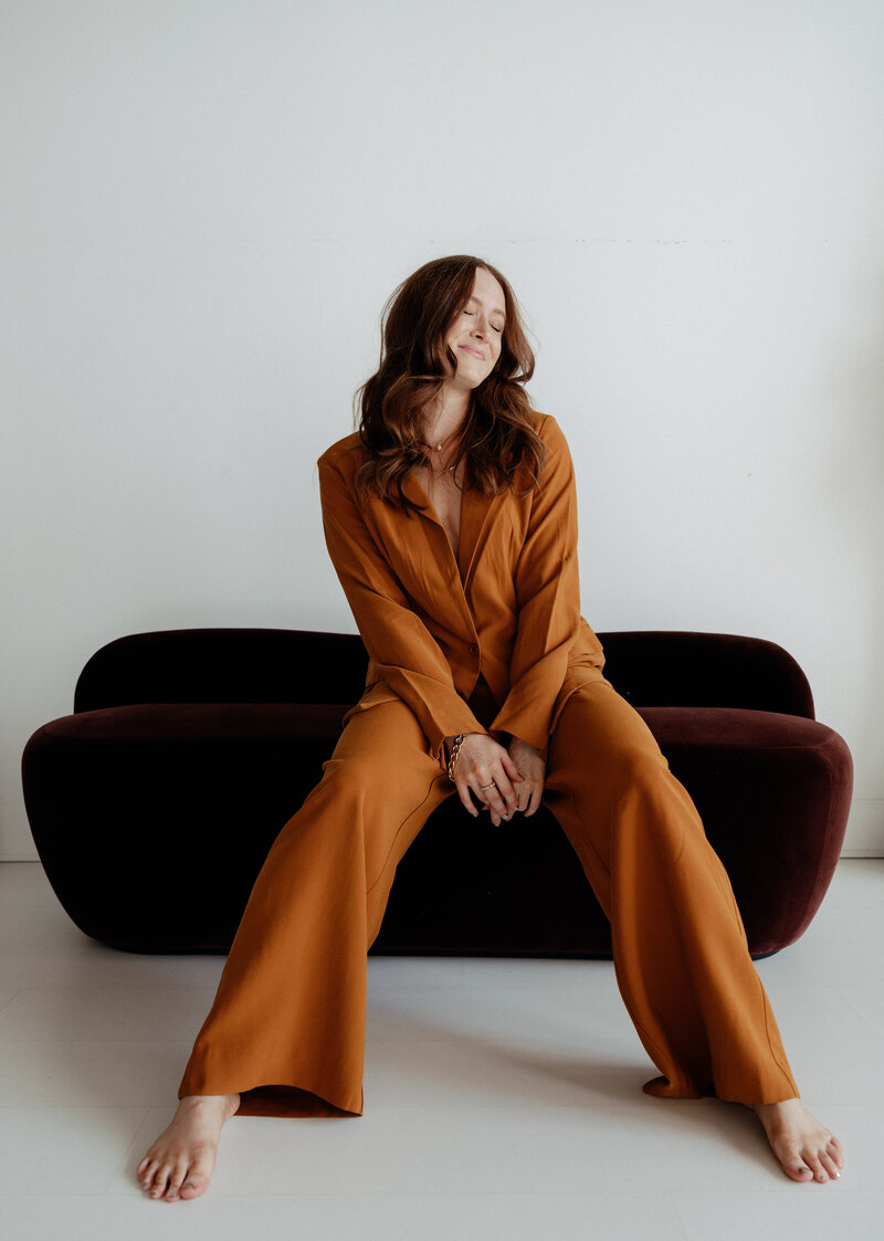 Atlanta-based lifestyle and brand photographer sitting barefoot on a modern velvet couch, wearing a stylish mustard suit and smiling with closed eyes.