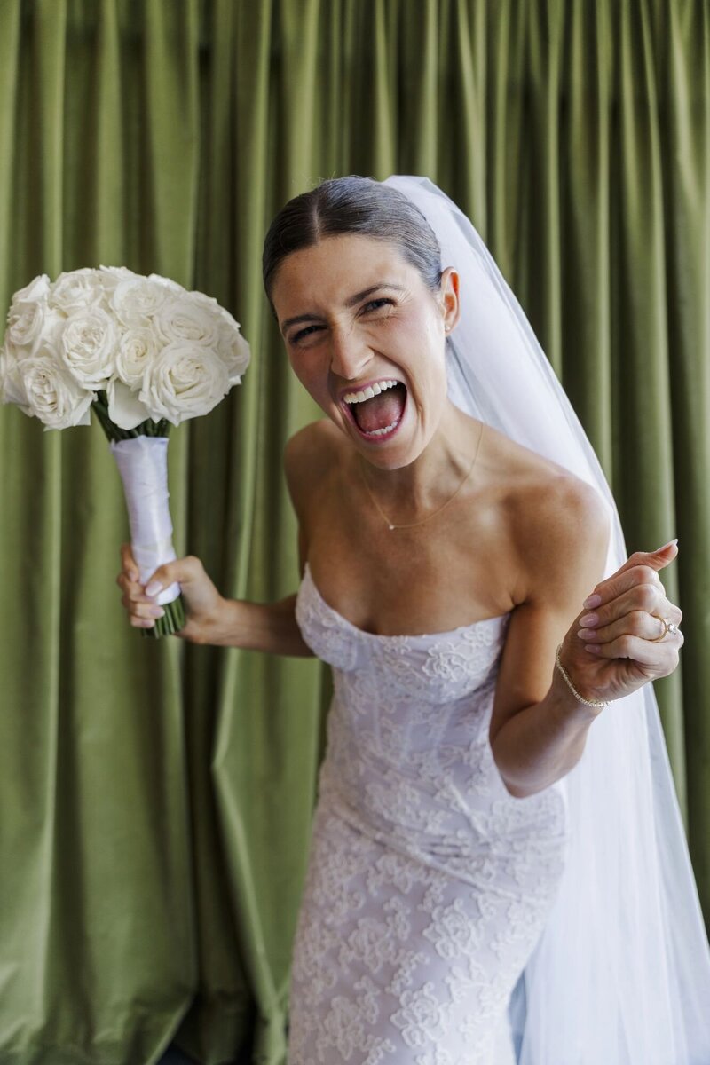 Bride in a corset gown cheering with a bouquet in front of a green velvet curtain