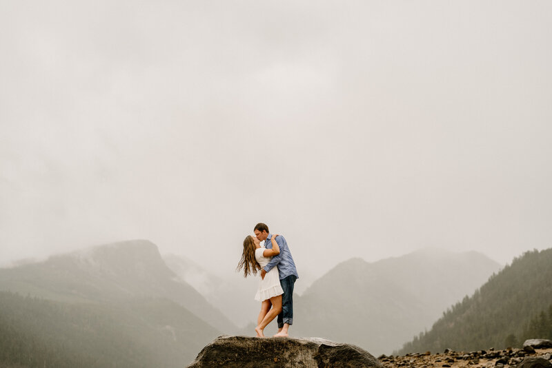 A couple kisses on a rock with mountains in the background