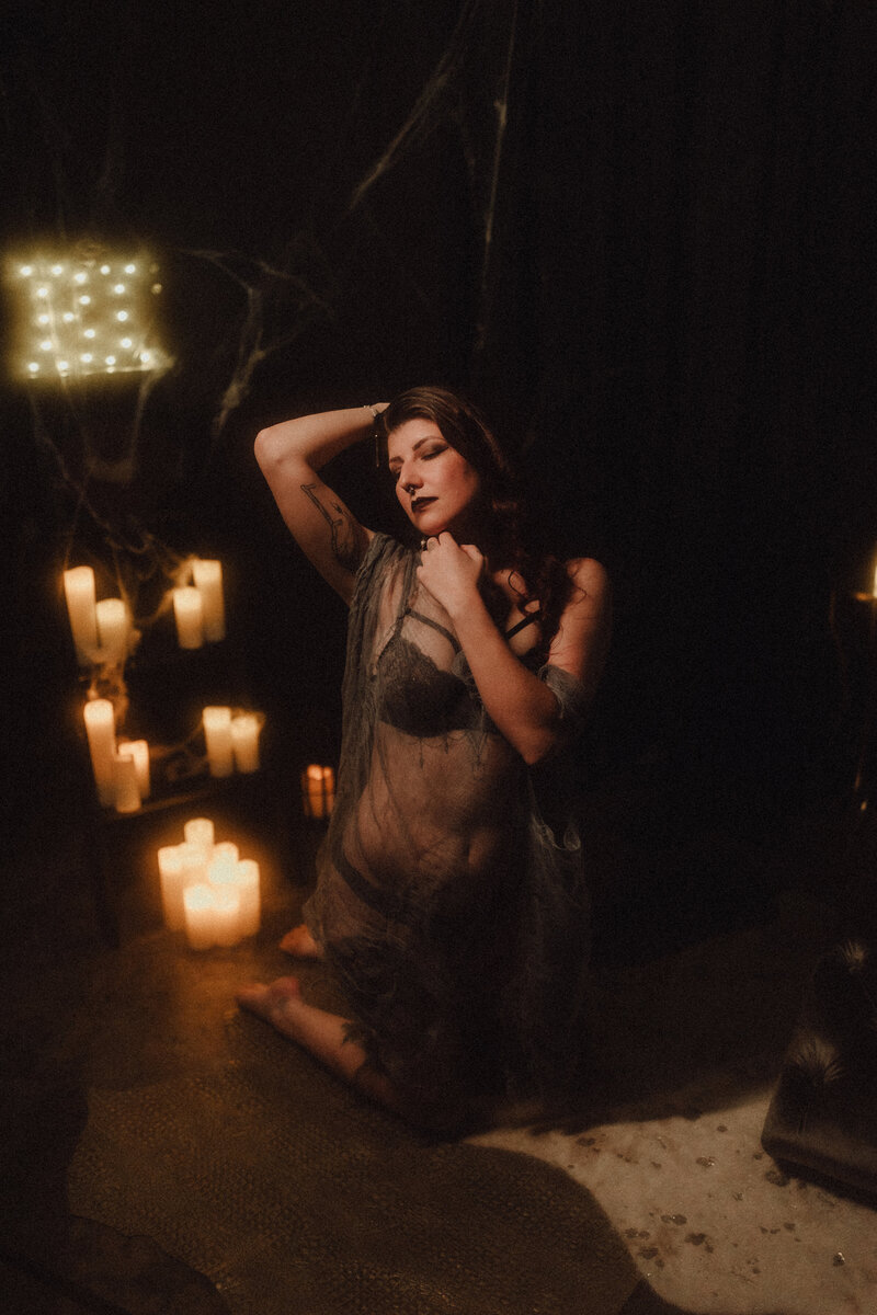 Tattooed woman in sheer robe poses gracefully by candlelight on velvet floor during dark glam boudoir session in Jacksonville.