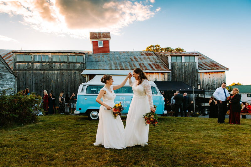 Two brides dancing in front of a vintage blue van at their outdoor New England wedding at Maple Rock Farm in Maine, holding colorful bouquets and smiling at each other.