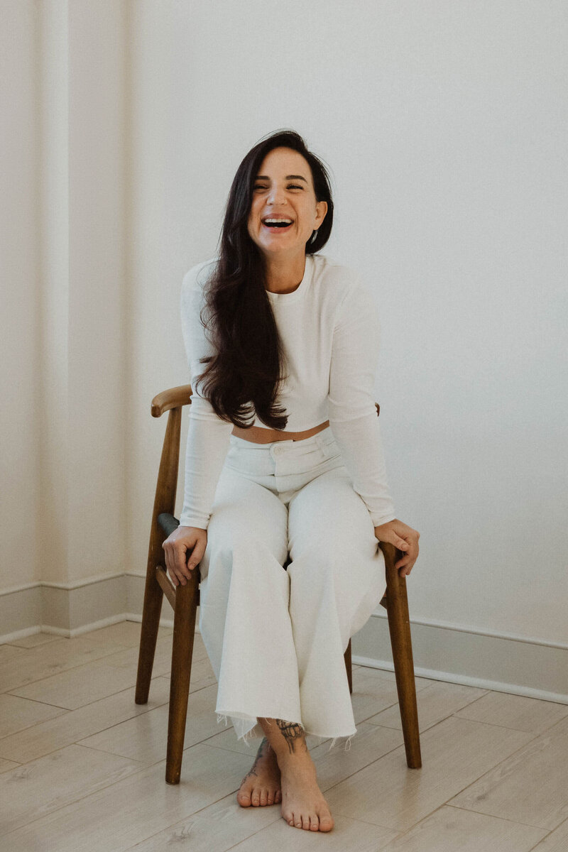 Ayurvedic skin specialist, Jen, sitting in a wooden chair smiling wearing a white top and pants