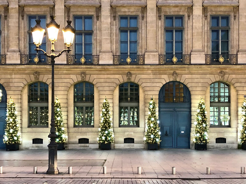 A historic building with elegant arched windows is adorned with a row of decorated Christmas trees. A classic streetlamp casts a warm glow, enhancing the festive atmosphere in the quiet, cobblestone street.