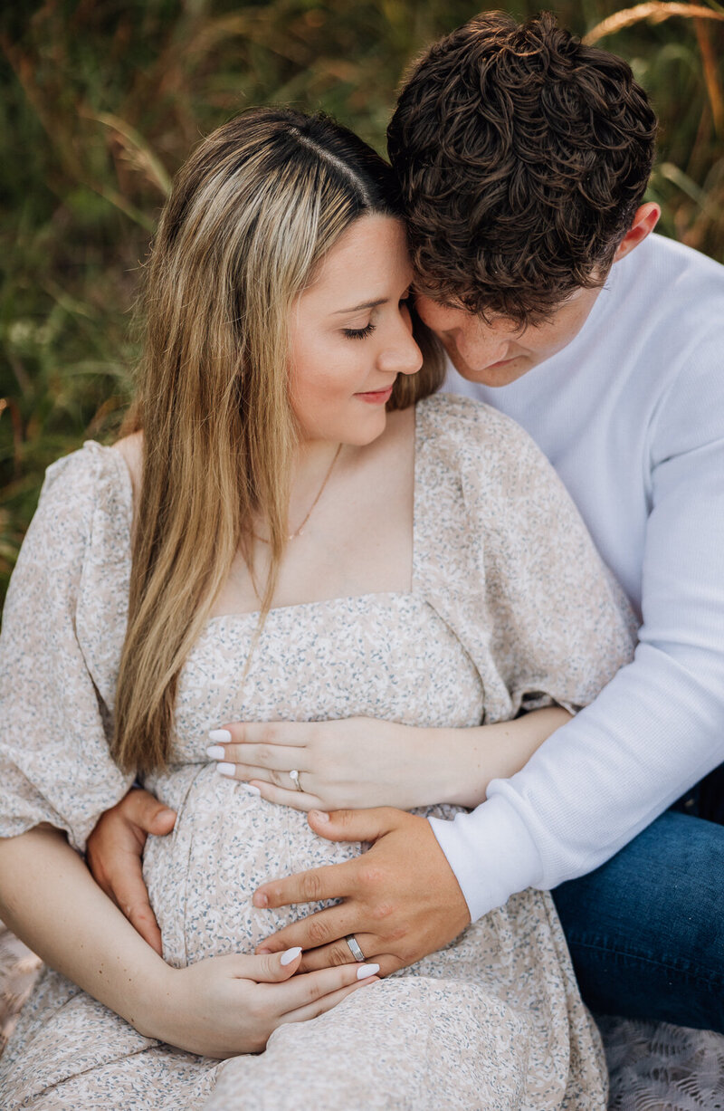 Lifestyle maternity photo of couple sitting on a blanket in a meadow, looking at each other lovingly.