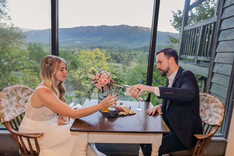 A bride and groom share a meal and wine after their elopement ceremony in celebration. They are sitting at a table with large glass windows overlooking a vast mountain range. 