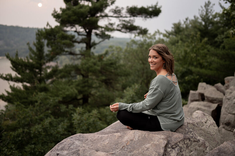 jen heller in a green shirt sitting on a large rock with a forest backdrop