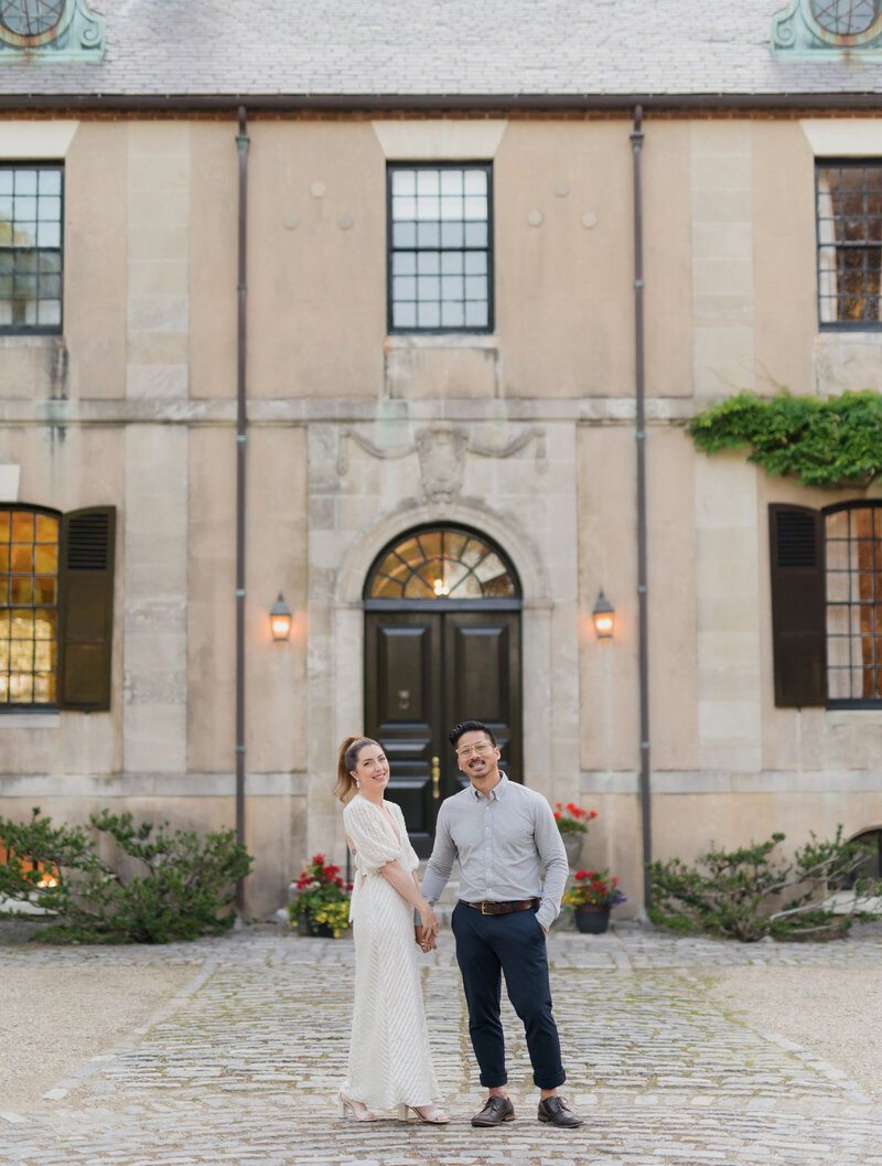 photographer and creative director kaylyn leighton holds husband hand in front of beautiful estate for an engagement session. Photographer, Joe sinthavong laughing at camera on a late summer day.