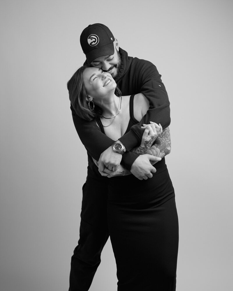 Black and white portrait of a couple embracing, radiating love and connection. The woman, wearing a fitted black dress and hoop earrings, smiles up at her partner as he wraps his arms around her from behind. He wears a hoodie and a baseball cap, his tattooed arms holding her close. A heartfelt, unfiltered moment from 'THIS IS ME – A Photography Experience by Katie Campbell' in Atlanta, GA, celebrating love and authenticity.