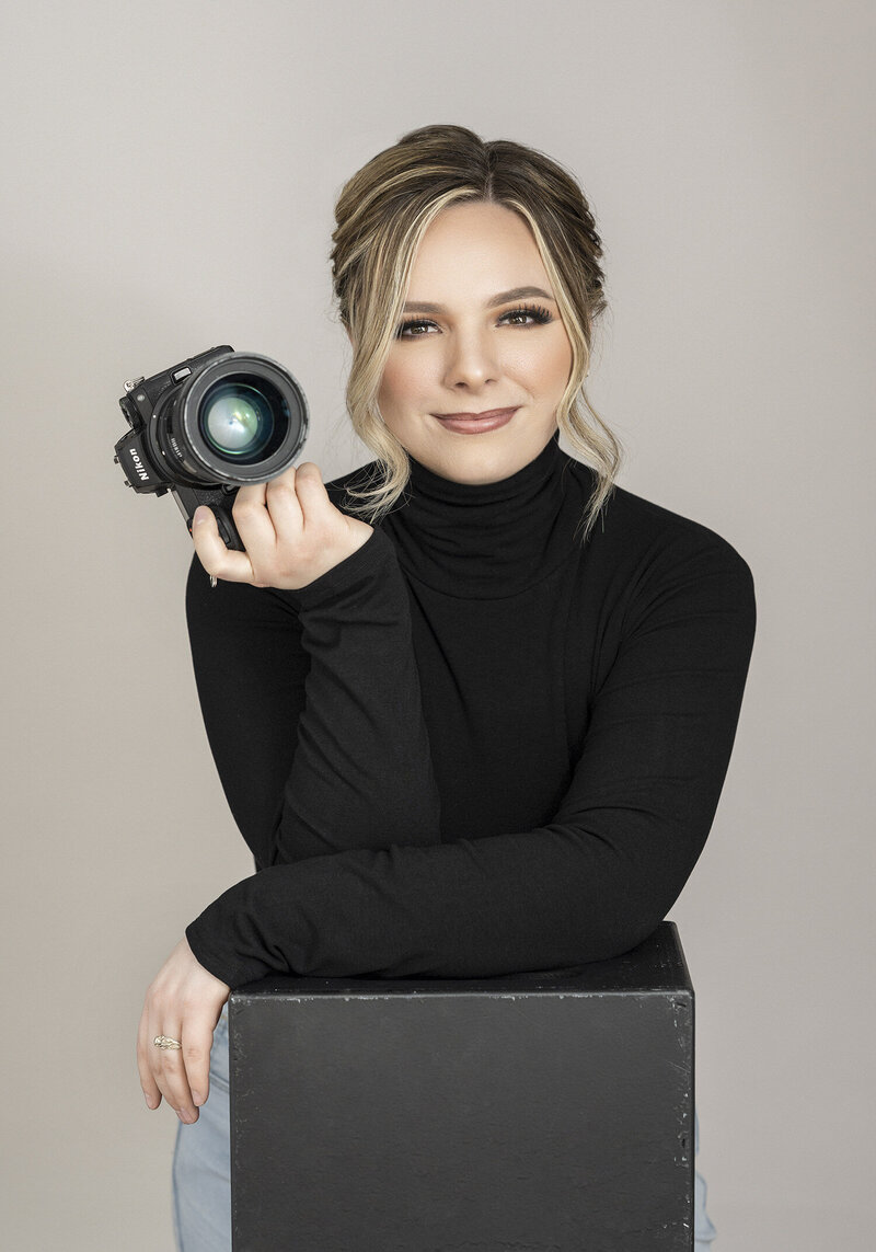 The owner of Kara Reese Photography poses in a black turtleneck with a Nikon camera in our Waukesha studio.
