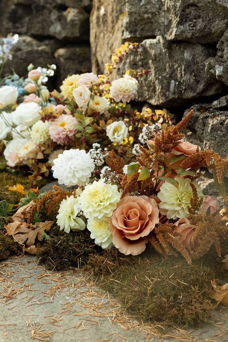 Close up photo of wedding ceremony flowers. The floral arrangements are on the ground with a rock wall behind them. They are sitting on top of moss. The flowers are bronze orange, cream, white, blush, and light blue.