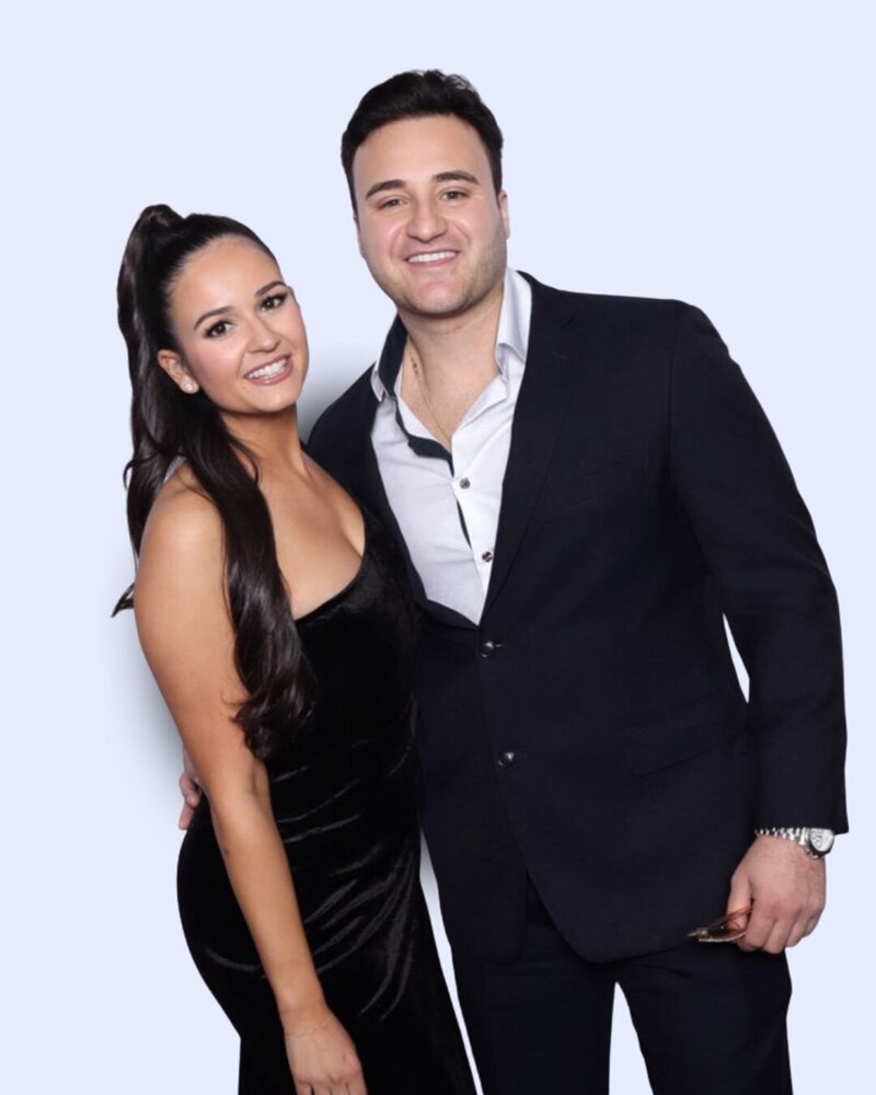 Couple dressed in a black velvet gown and navy suit posing together in front of a light-colored backdrop at AR Photobooth.
