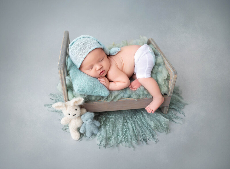 Newborn baby boy laying on a grey bed wearing white pants and a sleepy hat  taking first pictures at Rochel Konik  studio