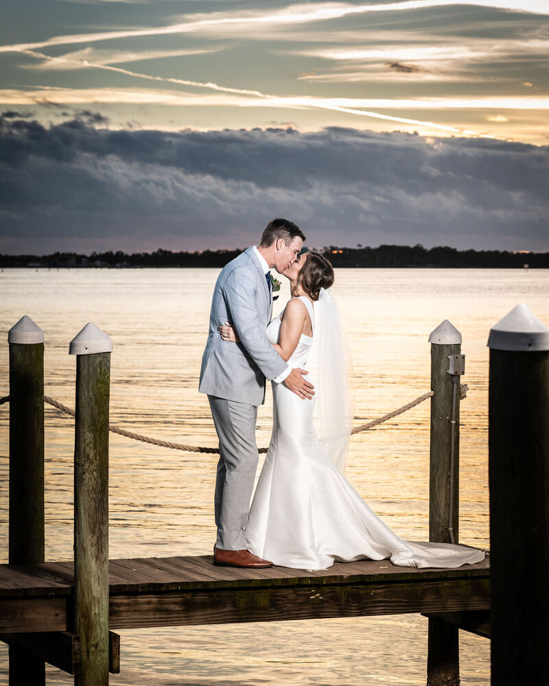 Bride and groom kiss on dock with sunset in background