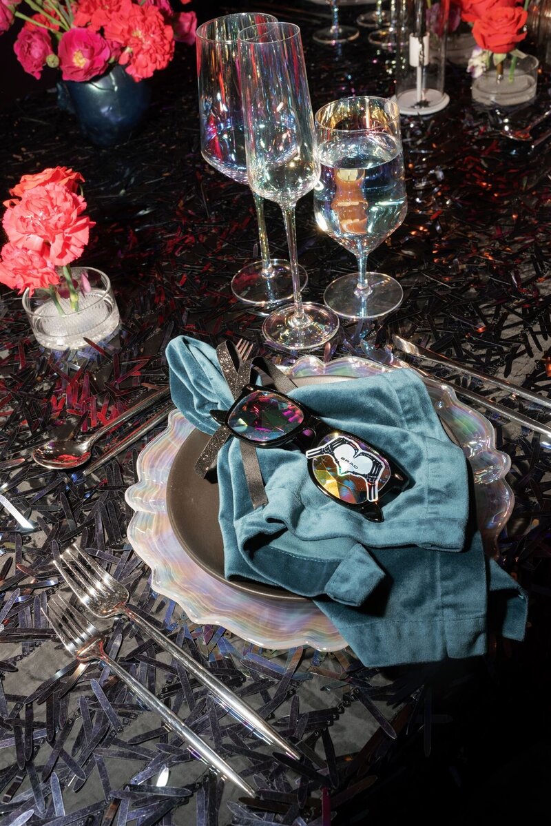 halloween meets disco inspired tablescape with black linen and multicolor sequins, silver flatware, iridescent glassware, iridescent charger with a black dinner plate on top,. a teal velvet napkin on top of the black plate, and black sunglasses with a skull heart place card sticker on top of the napkin