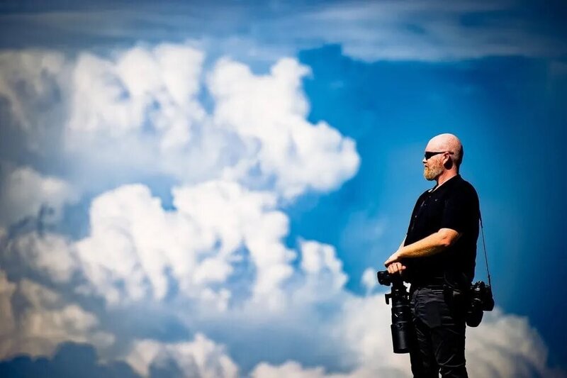 A photographer with a bald head and black shirt stands with his profile to the camera, holding two cameras against a vibrant blue sky with fluffy white clouds.