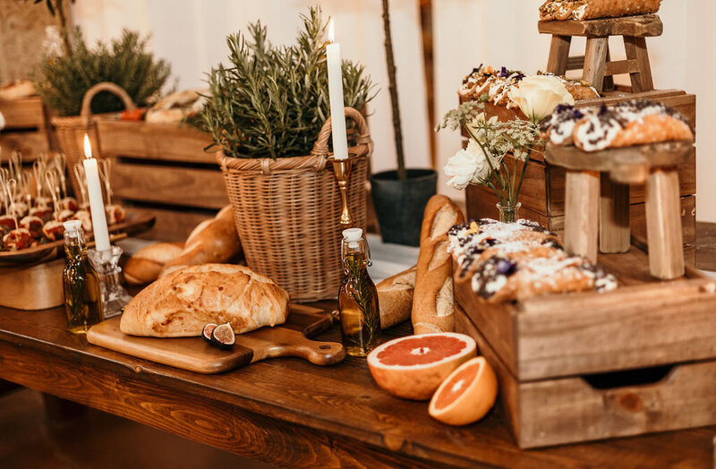 Wooden farmhouse table used as a grazing station featuring rustic bread, olive oil bottles, fresh fruit, and woven baskets.