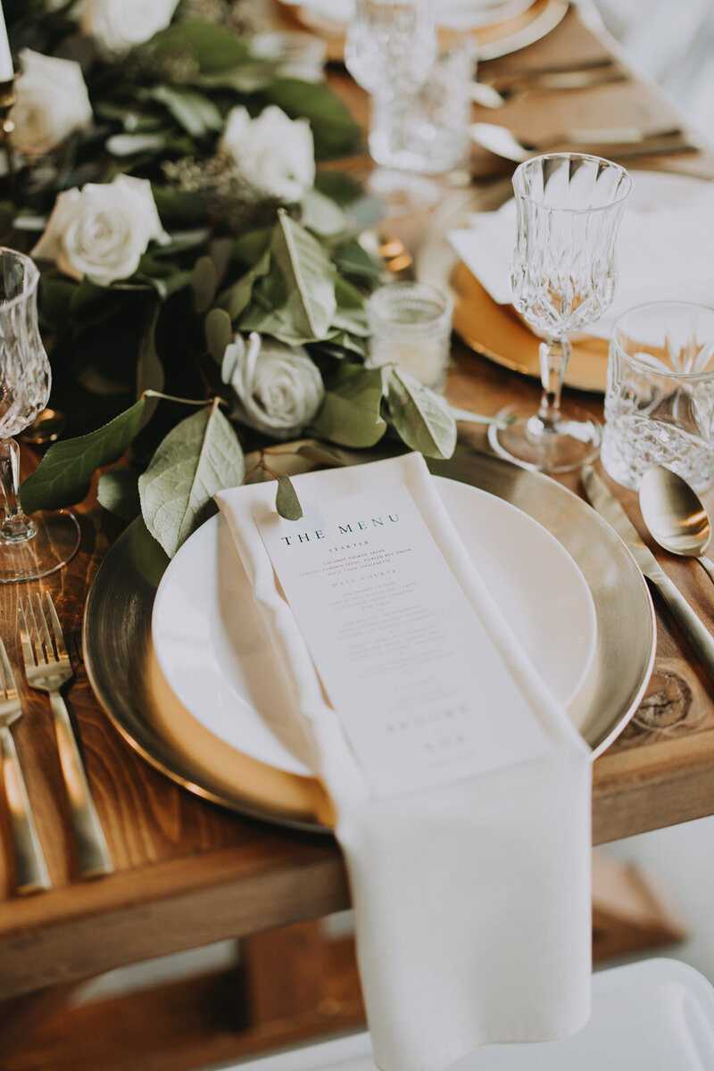 Close-up of a luxury wedding place setting with a gold charger, modern flatware, and crystal stemware surrounded by greenery.