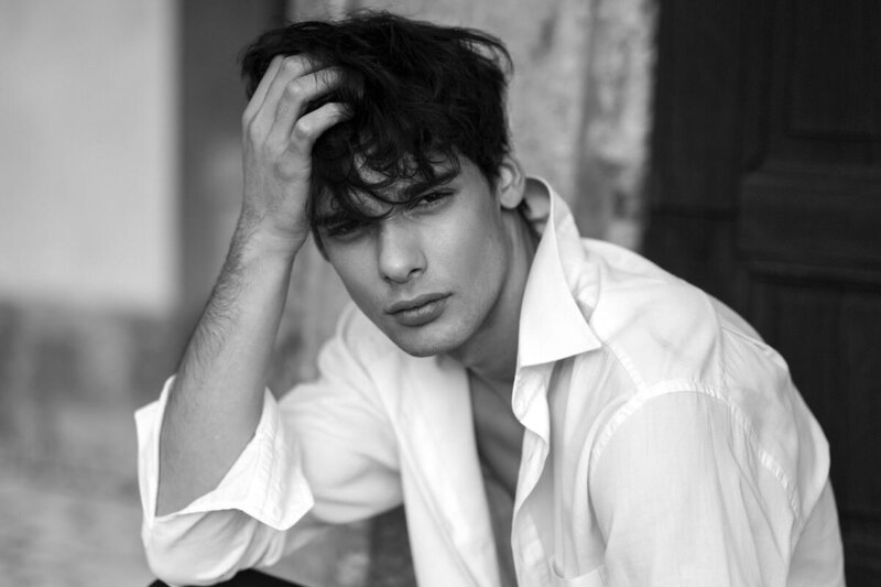 A black-and-white portrait of a young man with tousled hair, wearing a white button-down shirt. 