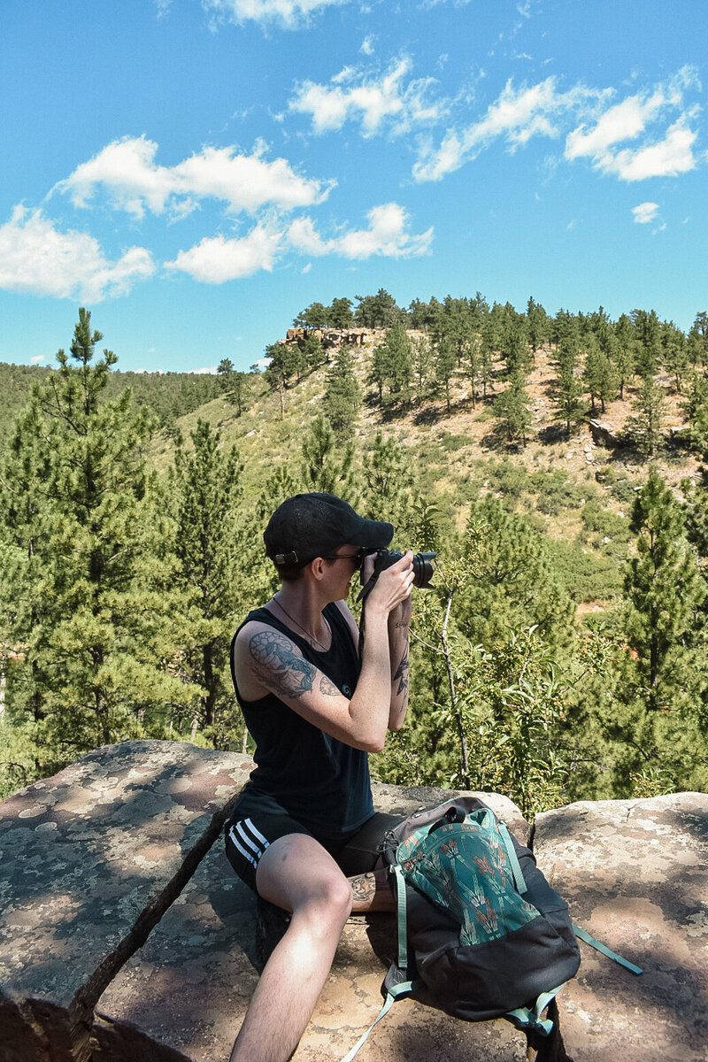 candid photo of photographer River Dantzler, who sits on a rocky overlook using their camera to take a photo. pine trees in the background.