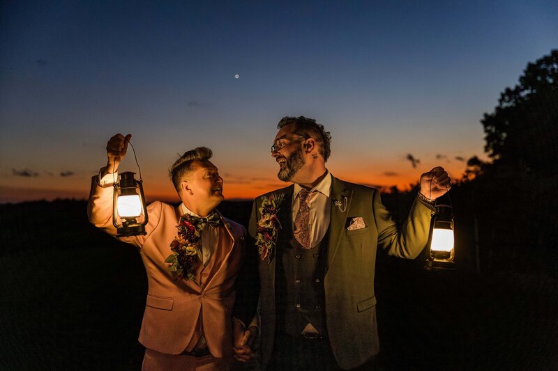 Two grooms look lovingly at each other while smiling and laughing. They are walking holding hands while lighting the way with lanterns. They are walking through grape vines at a vineyard after eloping. The sun has set behind them turning the sky bright orange. 