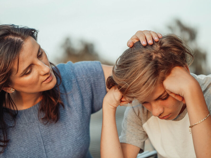 A mother and son sit outside together, the teenager resting his face in his hands. His mother gently strokes his hair with a caring expression on her face.