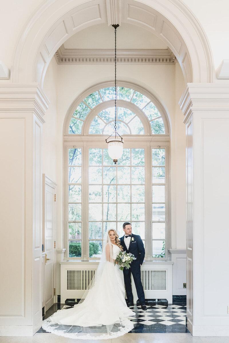 Bride and groom pose beneath an arched window with checkered flooring, bathed in natural light and classic architectural charm.