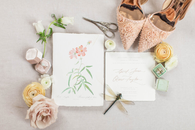 Wedding invite with flowers arranged around it and wedding details with pink shoes and a dragonfly