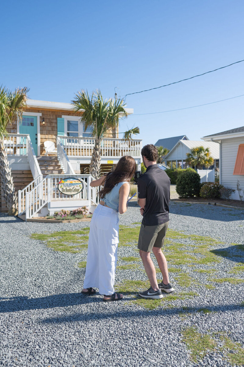 couple looks at the camera they're holding, backs to the camera with beach house in the background