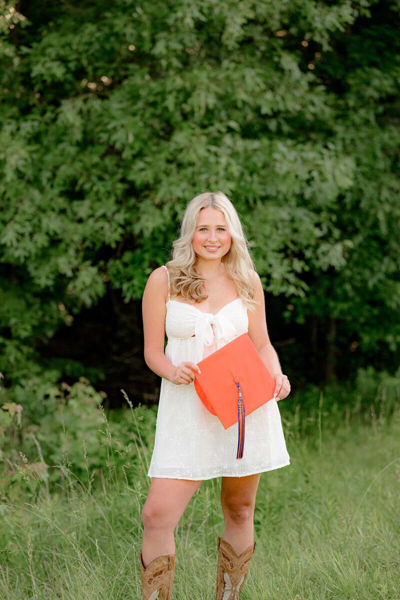 holding her orange graduation cap this MRHS senion poses in the tall grass while wearing a white dress