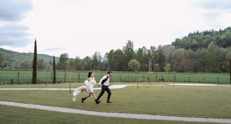 Bride and groom running hand in hand across a green field with mountains in the distance, captured in a joyful and cinematic wedding video set in rural Italy