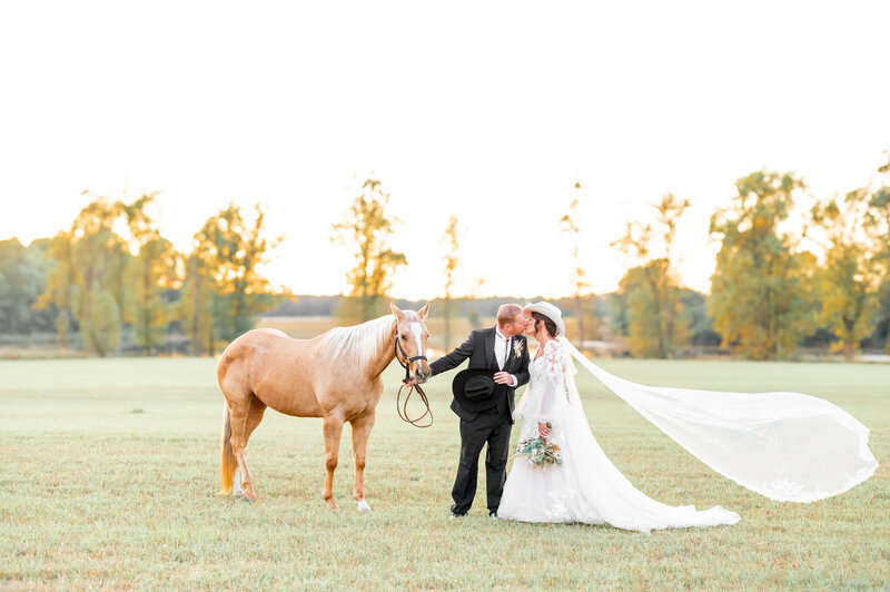 A bride and groom kissing in an outdoor grass field. The man is holding the reigns of a light brown horse.