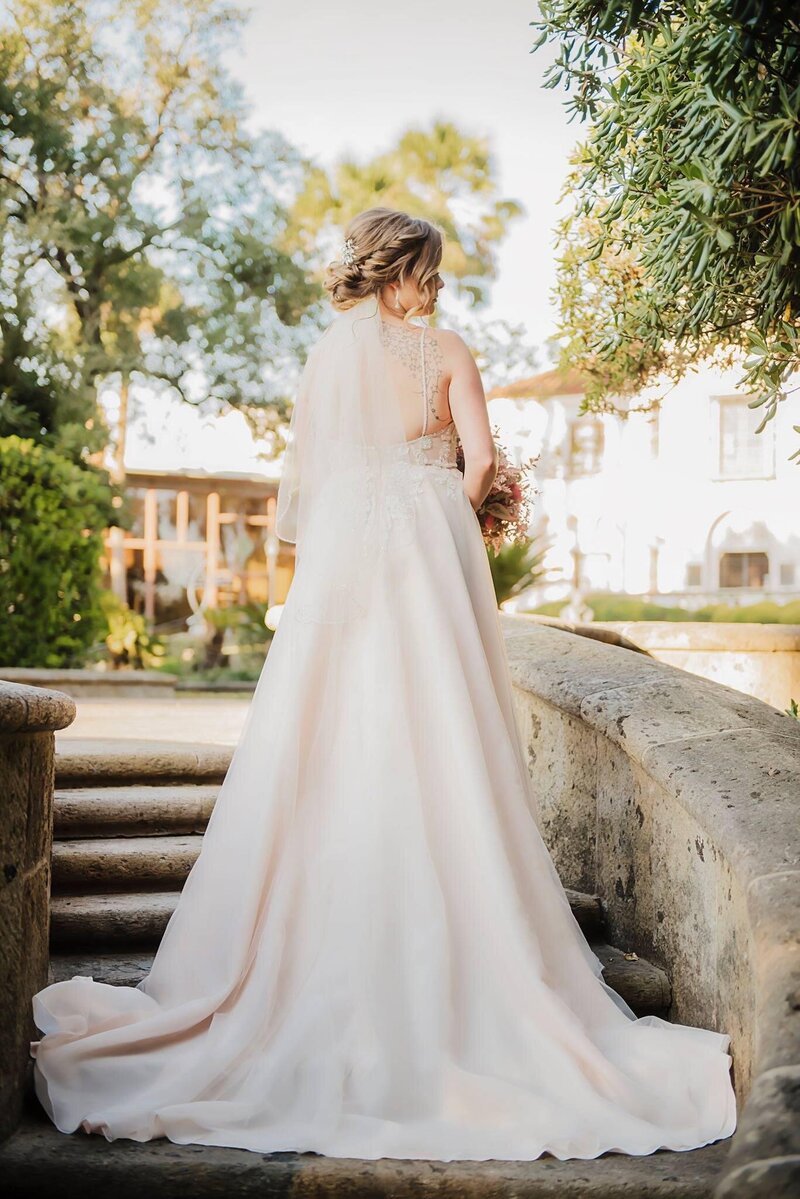 A bride in a white gown gracefully stands on stone steps, surrounded by lush greenery and a stately building in the background. Her elegant look is enhanced by one of Texas's finest wedding hairstylists, showcasing the stunning results achievable with premium bridal hair and makeup packages.
