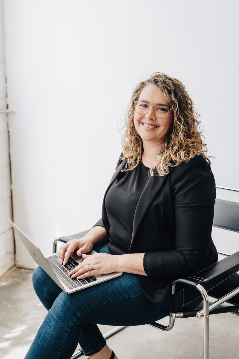 Woman sits with her computer on her lap smiling at the camera. 