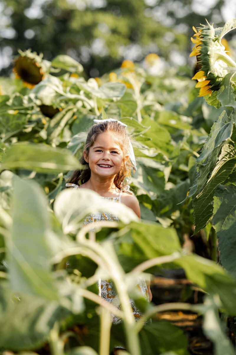 Young girl smiles while standing between tall sunflowers at sunset in Noblesville, Indiana