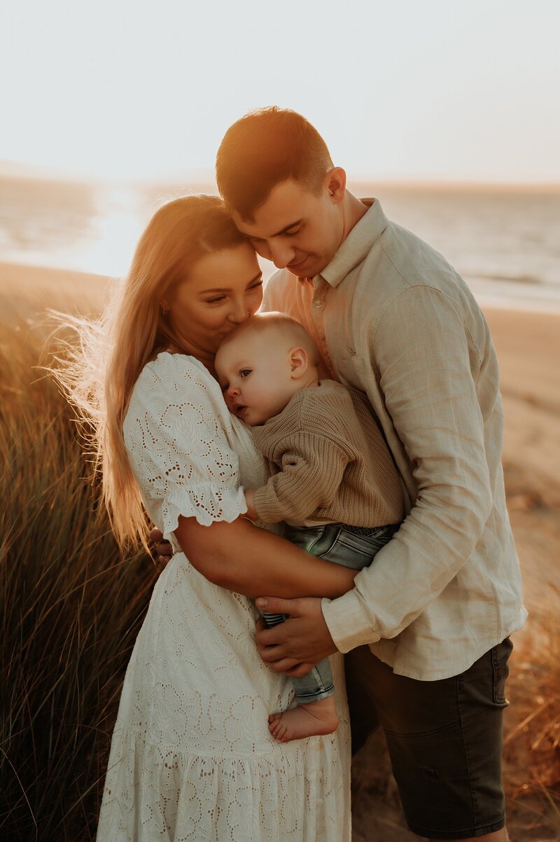Young couple holds their young child  between them as they hug. They are on the beach at sunset and having family photos taken