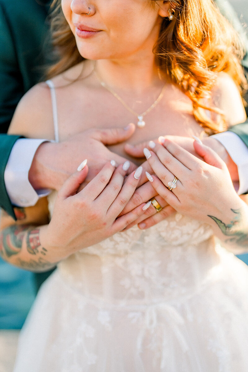 A close-up shot captures the heartfelt connection between the bride and groom during their California wedding. The bride, with soft waves and a delicate smile, holds her partner’s hands lovingly across her chest. Their rings sparkle in the warm California light, symbolizing the commitment they share. This intimate moment showcases the beauty of love and devotion, framed by the stunning details of the bride’s gown and delicate tattoos, highlighting the authenticity of this special day.