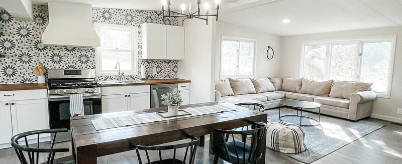 Explore a modern, open-concept kitchen and living area featuring a patterned backsplash and sleek white cabinets. The dining table with black chairs complements the light gray L-shaped sofa, while large windows fill the space with natural light. Visit our website for more details: https://halcyon-watson-1.showit.site/the-venue.