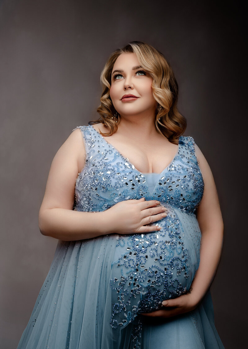 pregnant mother with hand on her shoulder on a brown backdrop wearing a cream sheer fabric 