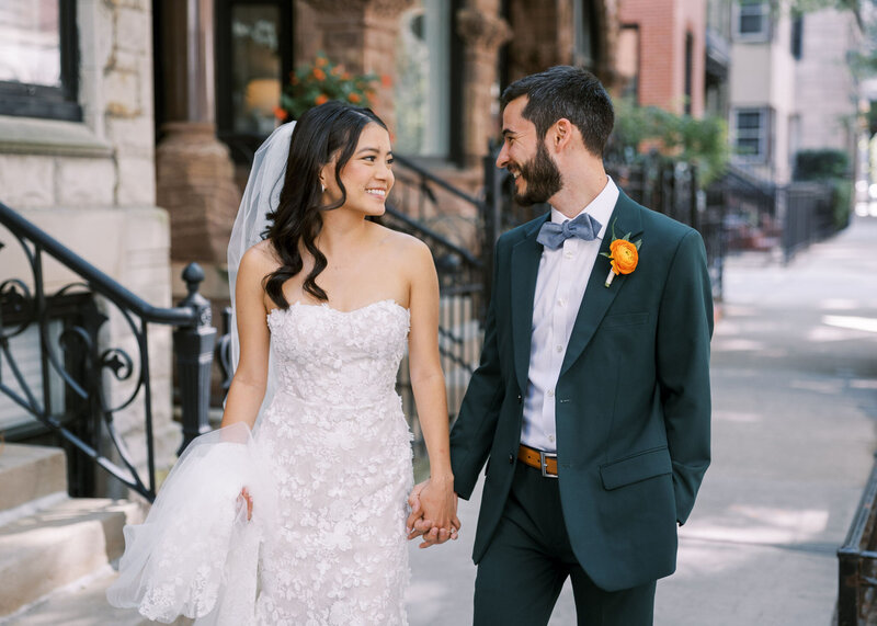 Bride in strapless flowery gown and veil hold's grooms hand as they smile at each other.