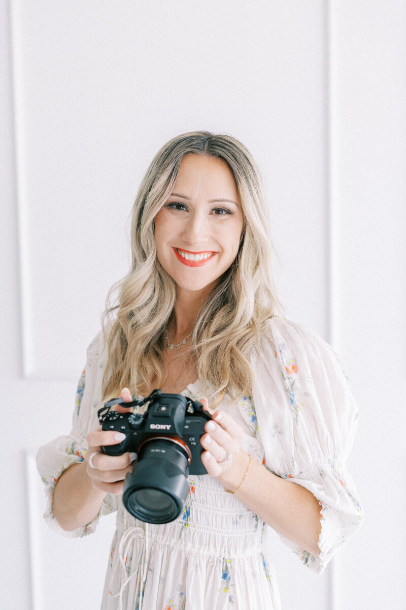 Nichole Pach wearing a white dress with flowers on it holding a sony camera and smiling 