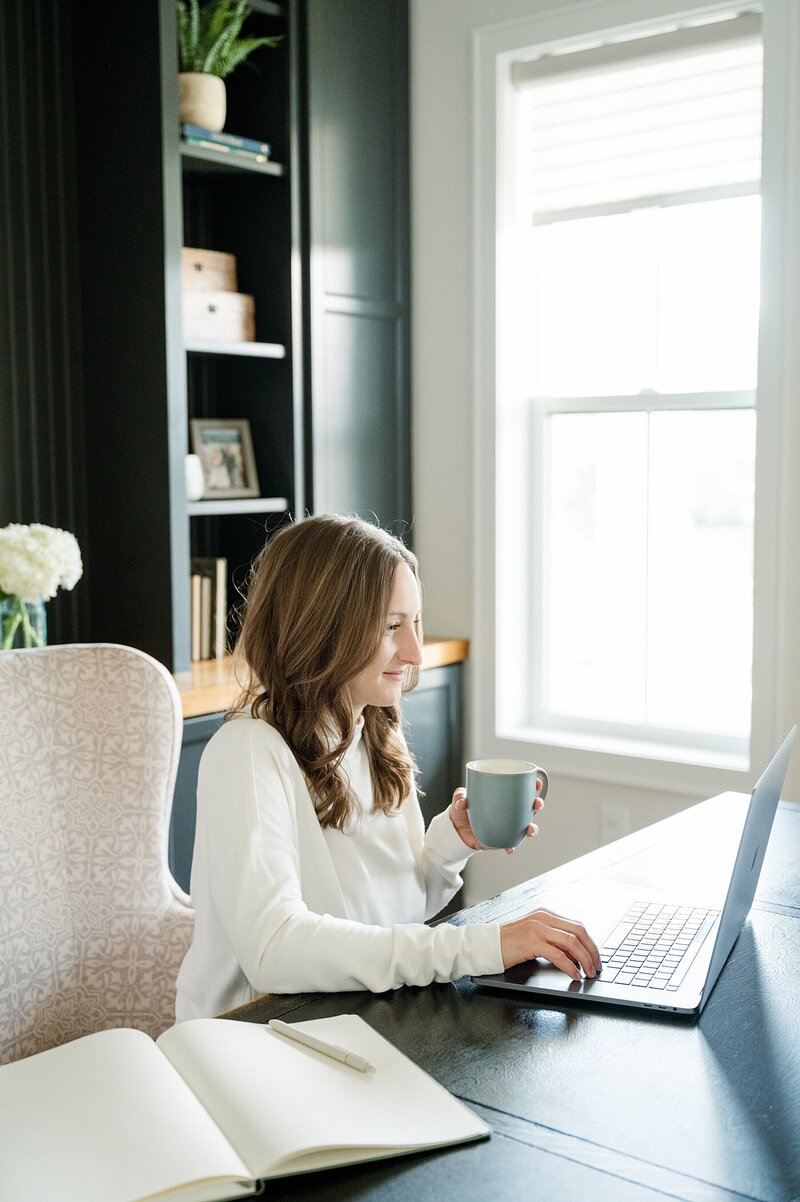 branding photoshoot with woman in a cream sweater sitting at her computer and holding a mug while she works and smiles for her branding photoshoot