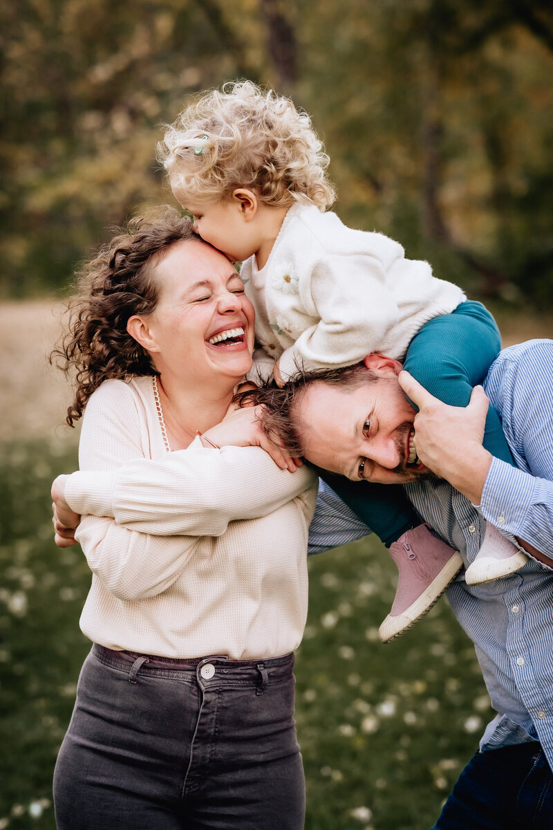 A joyful family moment captured outdoors, with a mother laughing as she carries her young child on her shoulders, while the father playfully leans in to join the fun. The child, perched on the mother's shoulders, is kissing her forehead.